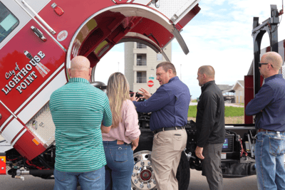 Ten-8 Fire & Safety representatives talk with fire department customers while pointing at an elevated Pierce fire truck cab and engine area.