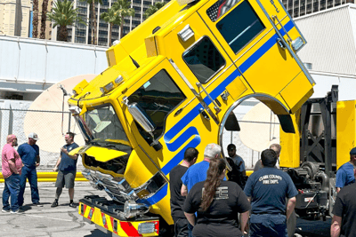 A yellow and blue fire apparatus with the cab tilted up as a group of firefighter's standby using the unit for training.