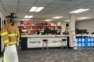 An indoor shot of a parts counter with two computer monitors and two stools. There is a mannequin dressed in a firefighter's jacket and helmet to the left. Shelves of parts are visible in the background.