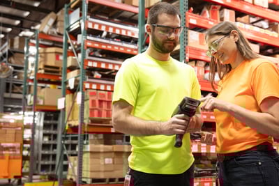 A man in a yellow shirt and a woman in an orange shirt look at a handheld scanner together standing in a warehouse in front of floor-to-ceiling shelves of boxes.