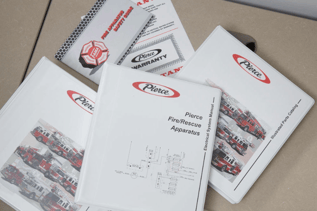 A top-down view of several binders and manuals from Pierce Manufacturing sitting on a desk. The titles on the binders include "Pierce Fire/Rescue Apparatus" and "Illustrated Parts Catalog."
