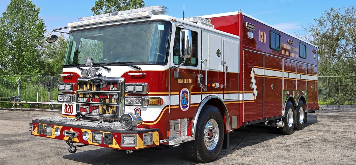 The red and white Prince George County Fire/EMS Department heavy-duty walk-in rescue unit is parked in a lot with the bumper in the foreground and trees in the background. 