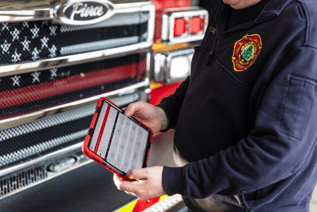 A man in a navy pullover with fire station emblem is standing in front of a fire truck and holding a tablet. The tablet screen displays diagnostic information.