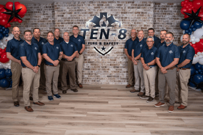 The Ten-8 Fire &amp; Safety team poses in matching blue shirts and khakis with the company logo behind them on a brick wall.