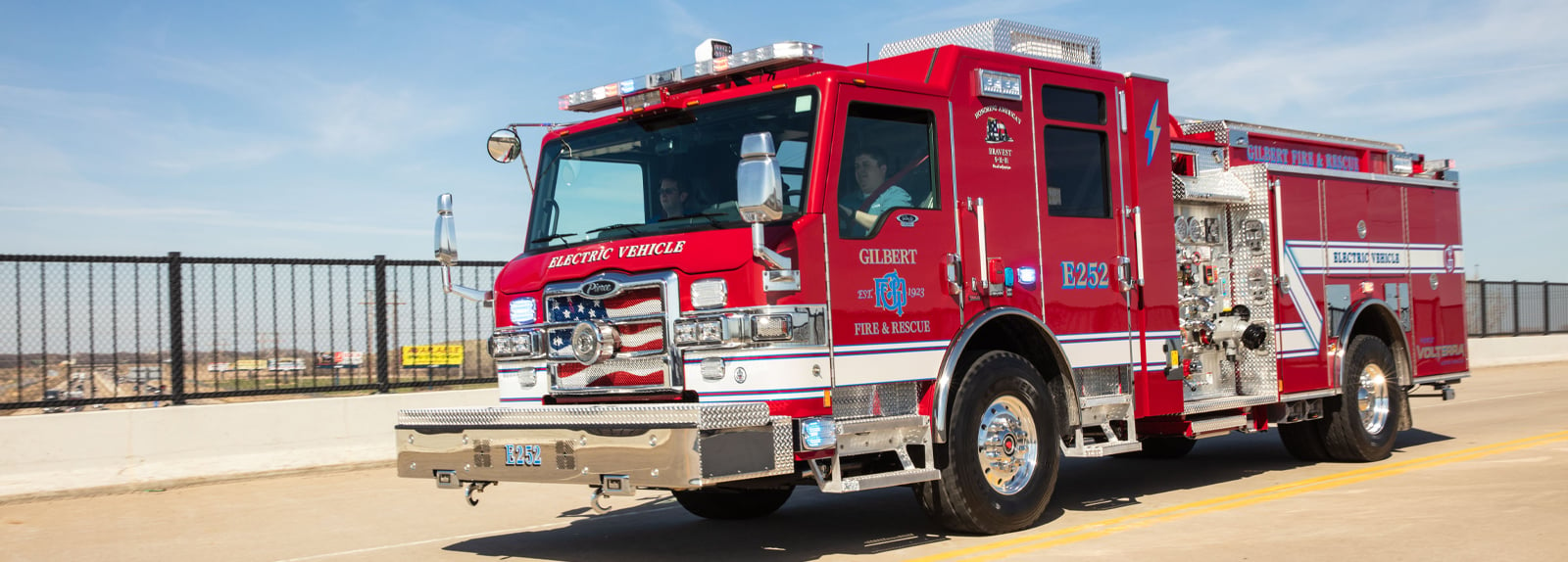 A red electric fire truck is driving on a roadway with a black rail barrier and blue sky in the background. 