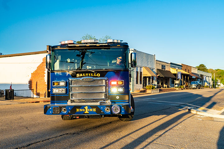 A blue Pierce Saber Pumper with its emergency lights on driving down a paved road in town.