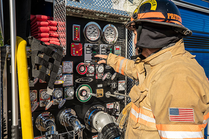 A firefighter in turnout gear operating a pump panel on a blue Pierce fire truck.