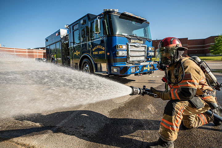 A firefighter kneeling in turnout gear spraying water from a hose in front of a blue Pierce Saber Pumper.