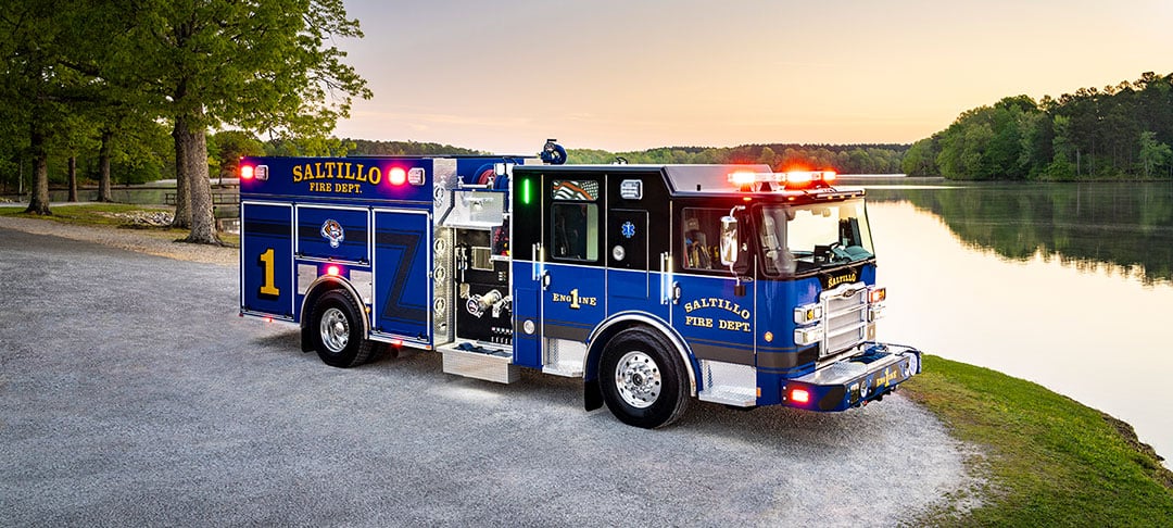 A blue Pierce Saber Pumper parked by a lake on gravel with sunset behind it.