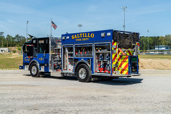 The driver's side rear of a blue Pierce Saber Pumper with compartments open showing gear parked in front of grass and an American flag.
