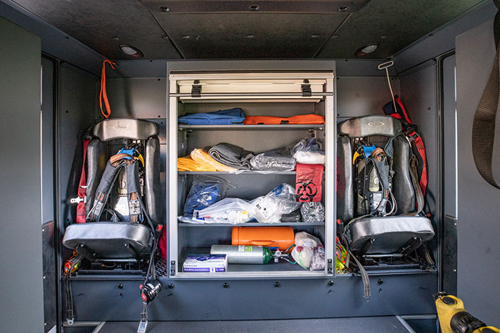 The inside of a crew cab of a fire truck with two seats and a roll-up compartment in the center that is open showing equipment.
