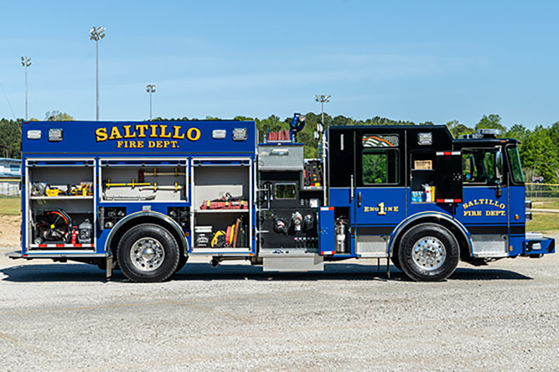 The officer's side of a Pierce Saber Pumper with compartments open showing equipment inside parked on gravel in front of green grass.