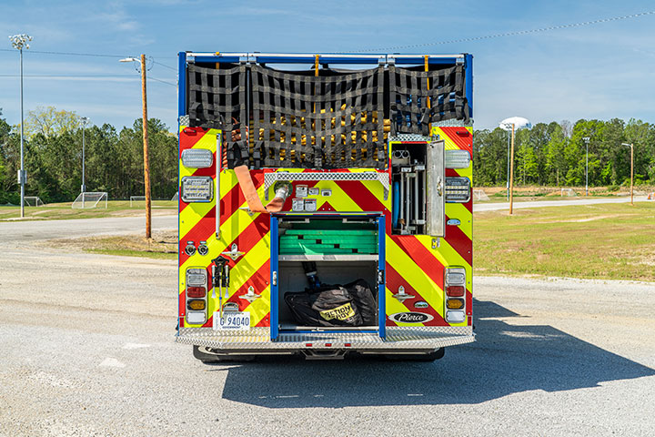 The rear of a Pierce Pumper with the compartments open showing equipment parked by green grass and trees.