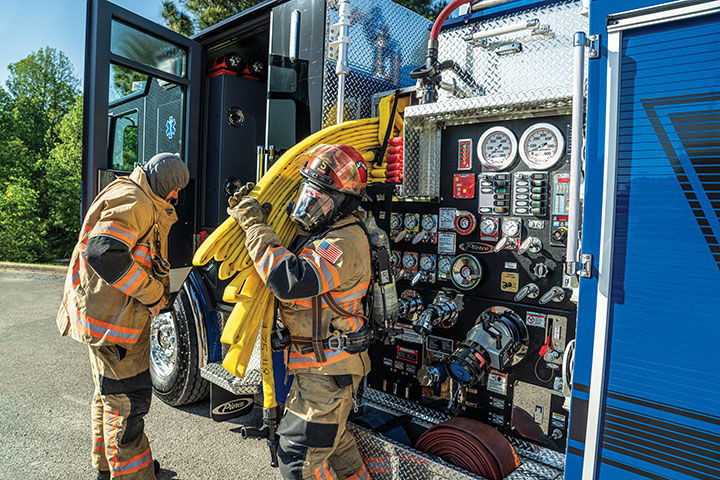Two firefighters in turnout gear next to a blue fire truck and one unloading hose from a crosslay by the pump panel.