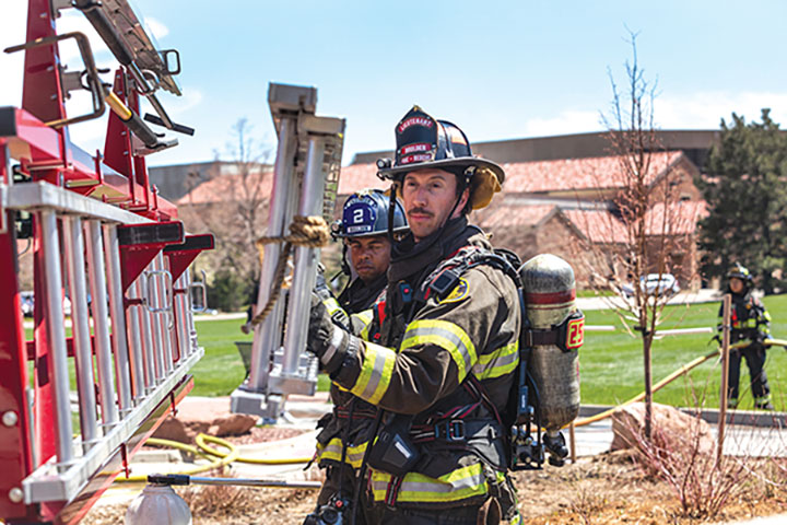 Fire fighter in gear taking a ladder off of the officers side of a fire truck outside with firefighters behind them.