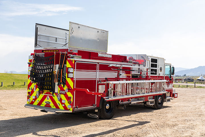 Rear of fire truck with top compartments open and hydraulic ladder down parked outside.