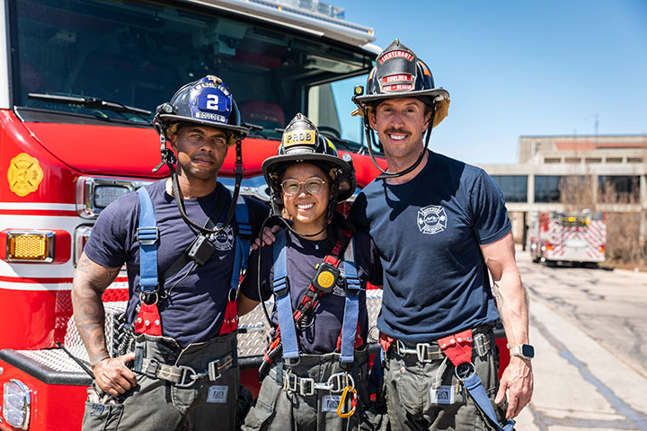 Three smiling firefighters in gear standing in front of a fire truck.