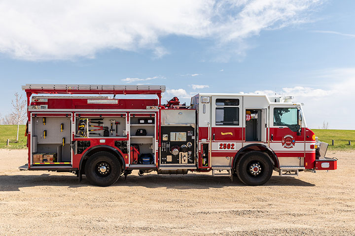 Velocity pumper officer's side of a fire truck with compartments open showing equipment parked next to a field of green grass.