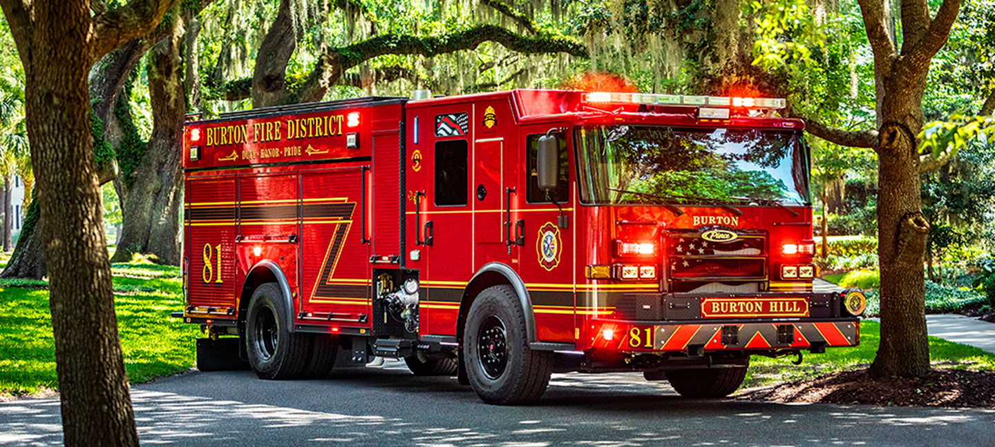 A Pierce Enforcer PUC Pumper parked under green trees on a paved road in front of green grass.