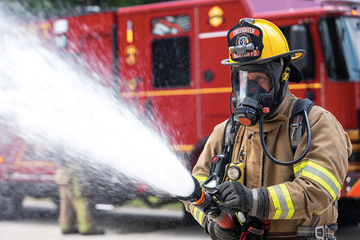 A Burton firefighter in turnout gear spraying water from a hose in front of a Pierce Enforcer Pumper on scene.