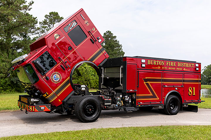 A Pierce Enforcer PUC Pumper with the cab tilted parked on a road by green grass and trees.