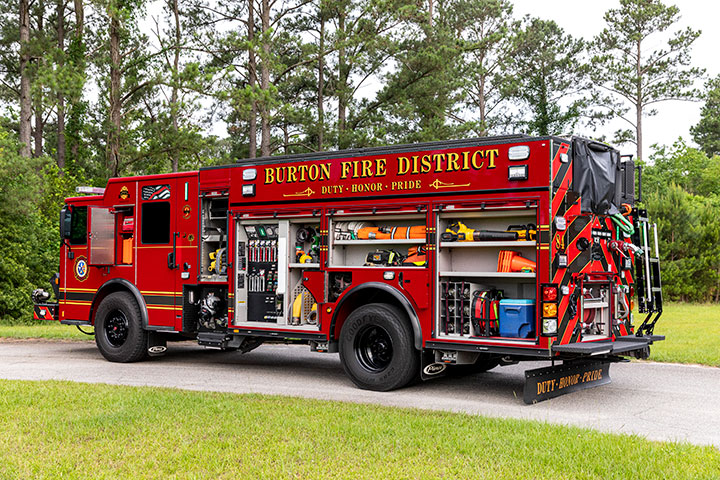 The driver's side of a Pierce PUC Pumper with the compartments open showing equipment parked on a paved road by grass and trees.