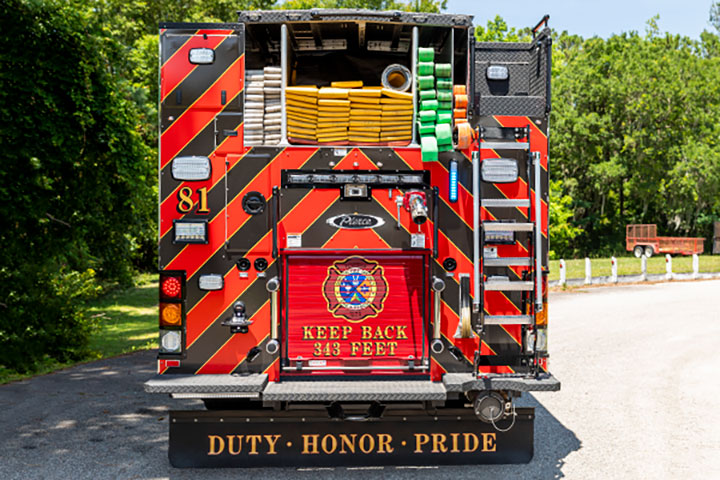 The rear of a Pierce Pumper with hose loaded in the hosebed parked next to green trees.