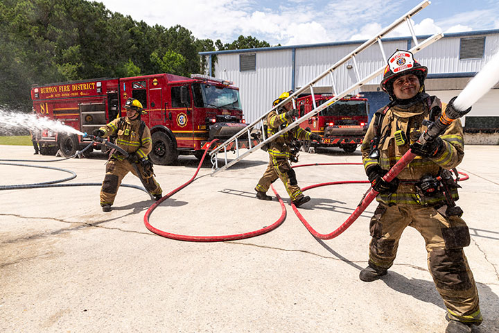 Three firefighters working on scene spraying water from hoses and carrying a ladder in front of a Pierce Enforcer PUC Pumper.