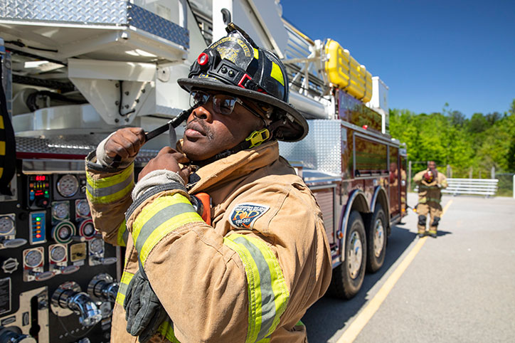 A firefighter in turnout gear standing next to the pump panel of an Aerial MidMount Tower putting his helment on.