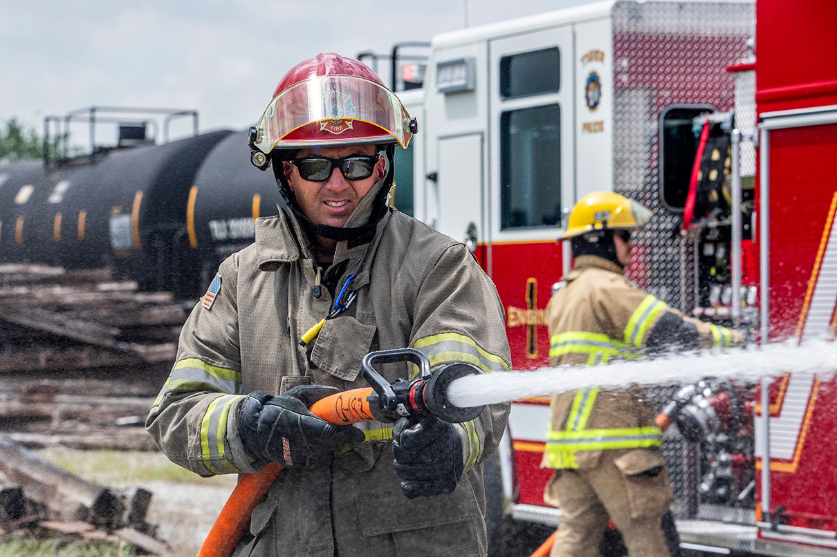 A firefighter in turnout gear holding a hose next to a fire truck with a firefighter at the pump panel beside a railroad train.