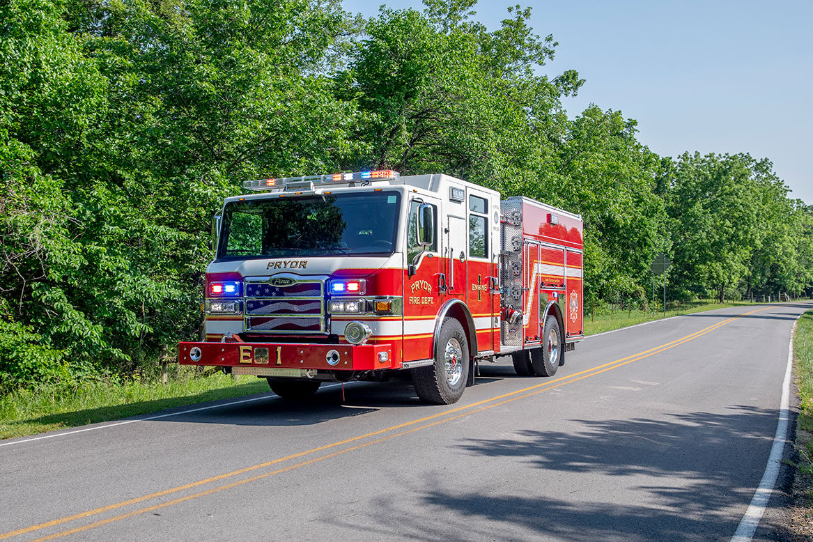 The front drivers side of a Velocity Pumper driving on a road next to a treeline on a sunny day with the lights on.
