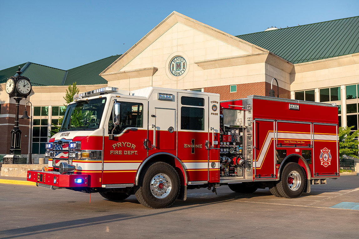 The front drivers side of a Velocity Pumper with the lights on driving out of a fire station on a sunny day.