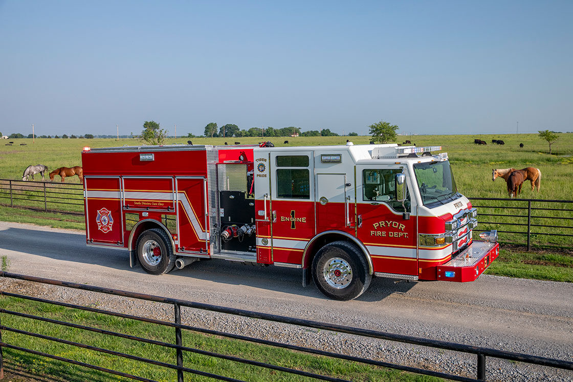 The front officers side of a Velocity Pumper with the lights on driving on a gravel road next to a fenced in pasture with horses.