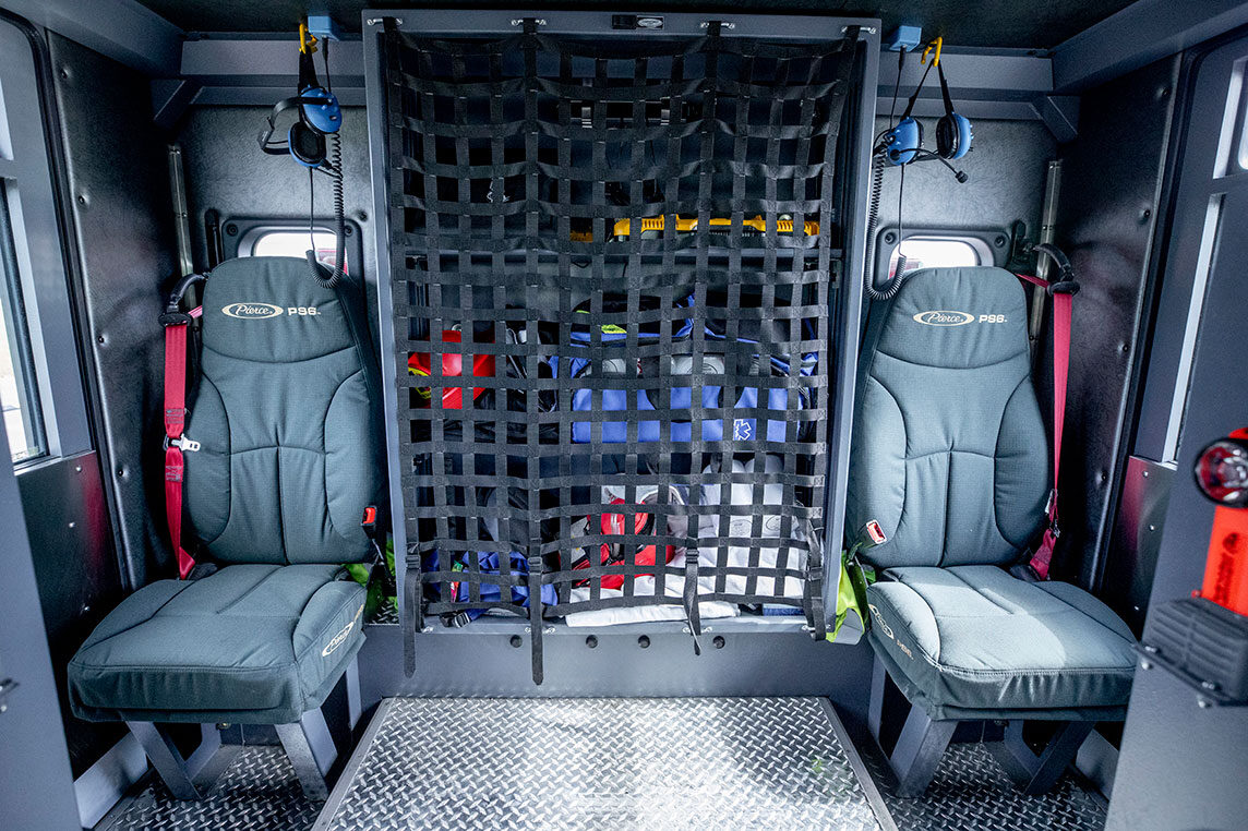 The inside crew cab of a fire truck with two forward-facing seats and a compartment holding tools and gear in the middle.
