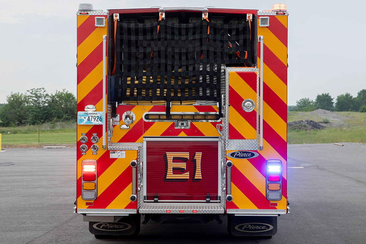 The rear of a fire truck with the lights on showing hose in the hosebed with a net hosebed cover.
