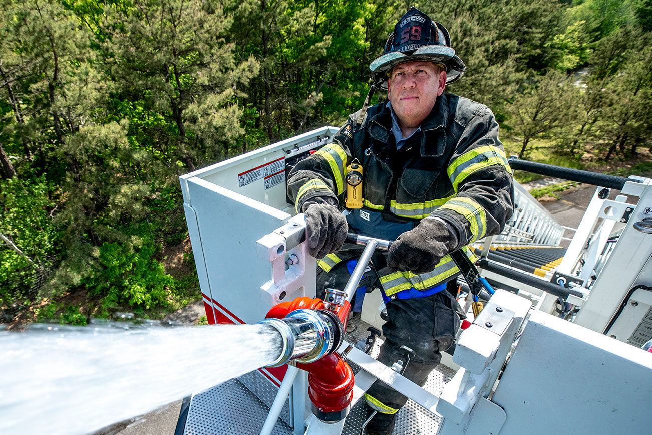A firefighter in turnout gear standing on elevated aerial platform bucket spraying water.