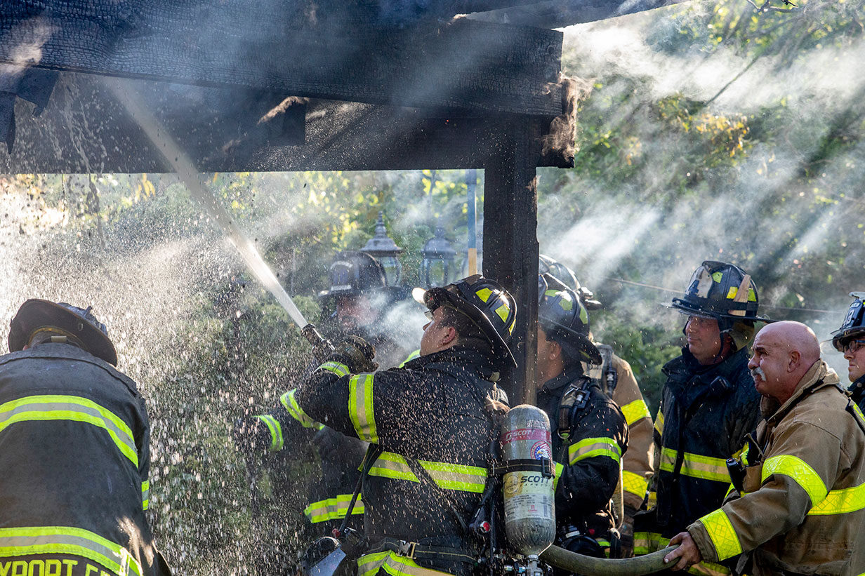A group of firefighters in turnout gear fighting a fire on a patio with a fire hose spraying water.
