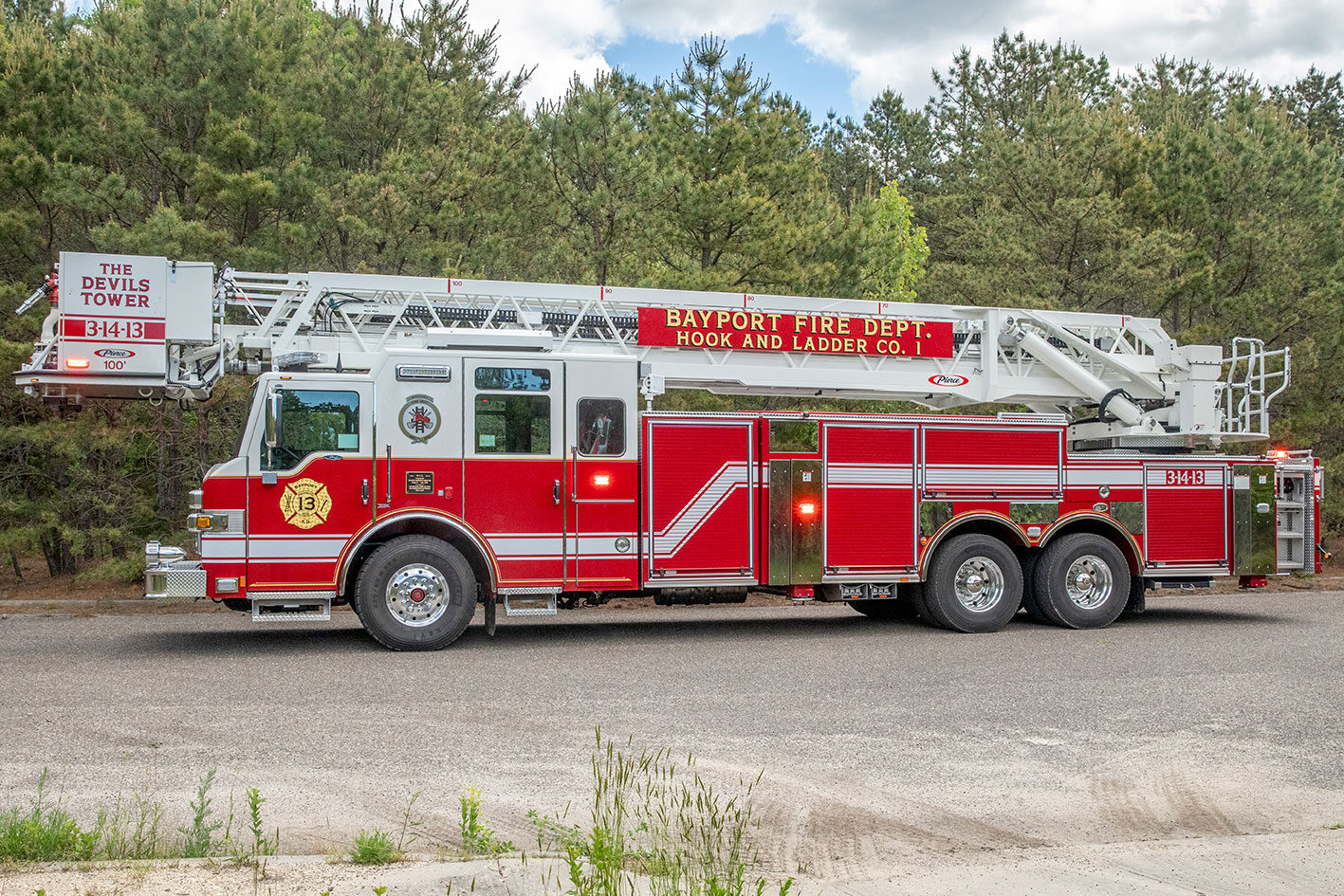 Th drivers side of an aerial platform fire truck with the lights on parked on a road next to woods.