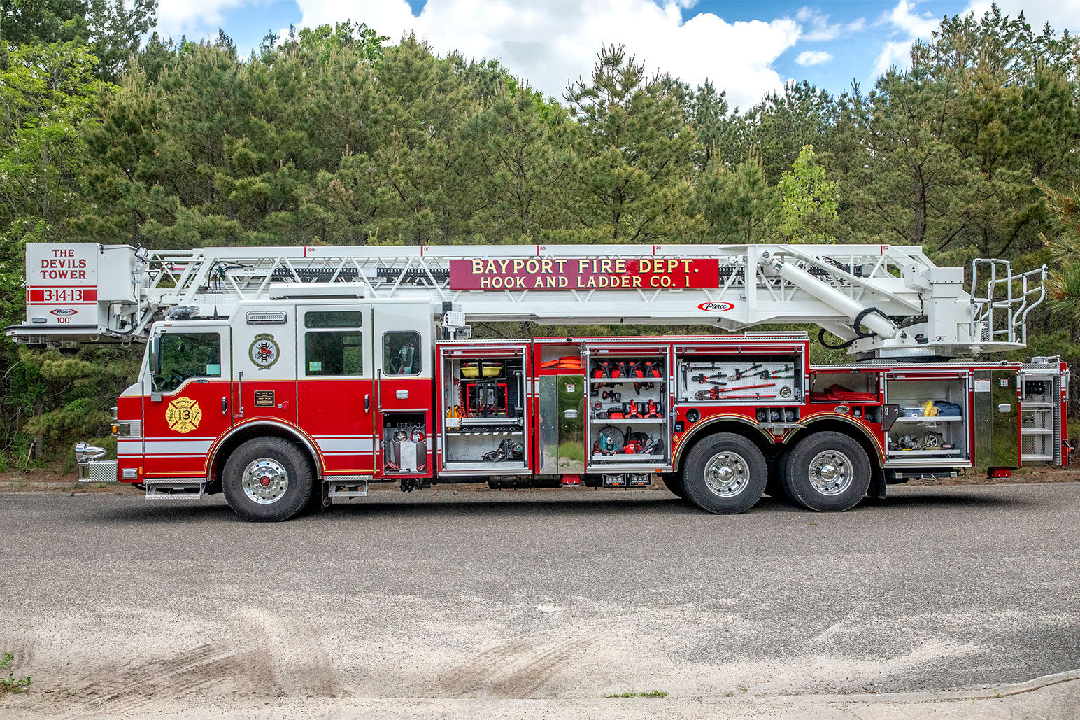 The drivers side of a aerial platform fire truck with the compartments open showing equipment and tools inside next to the woods.