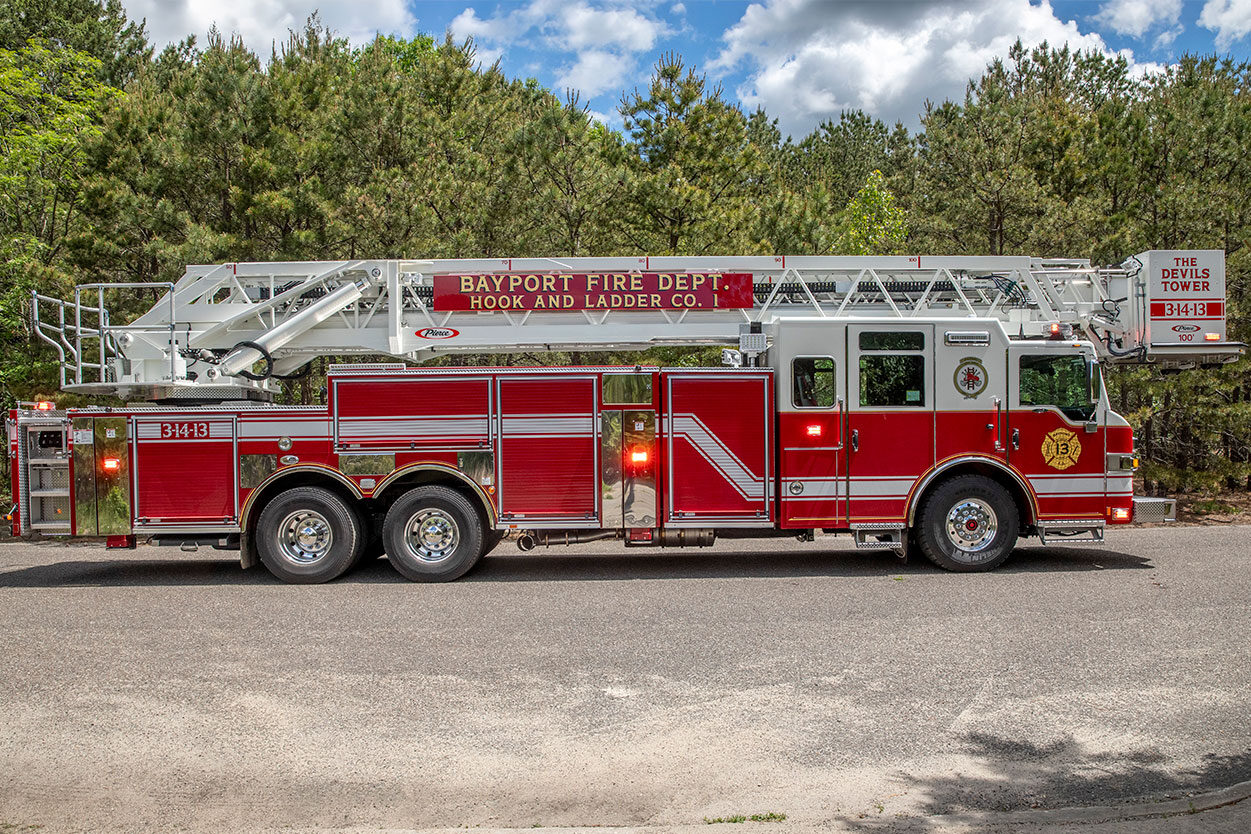 The drivers side of an aerial platform fire truck with the lights on parked next to the woods.