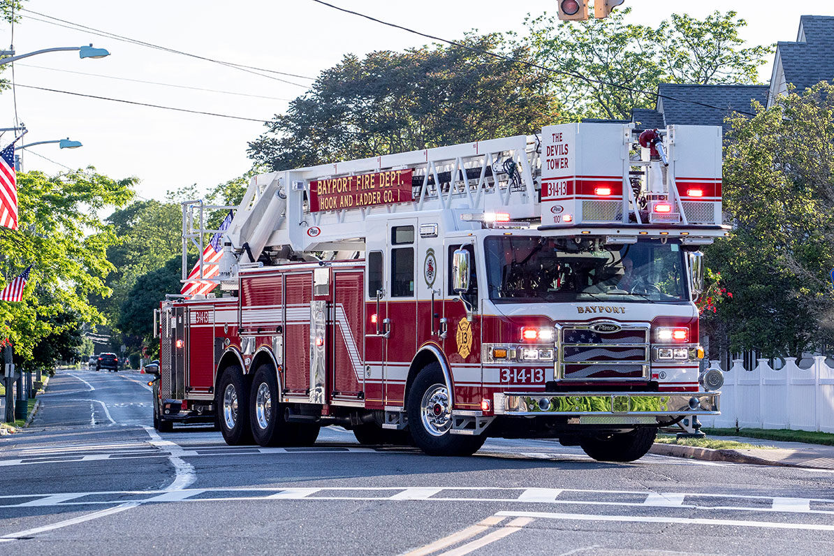 The front officer's side of an aerial platform fire truck driving down a street with its lights on.