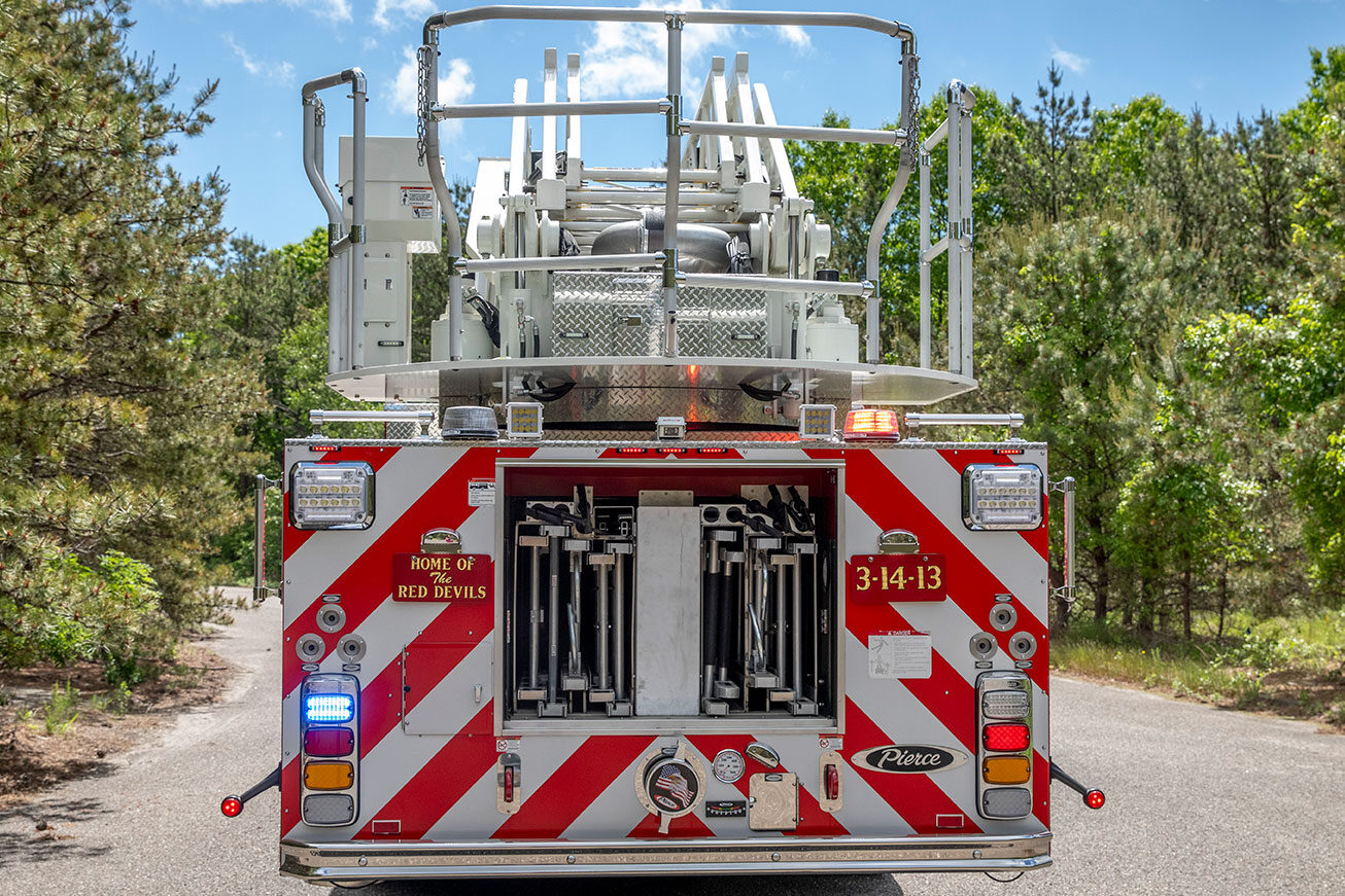 The rear of an aerial fire truck parked next to the woods with the compartment open showing ground ladder storage.