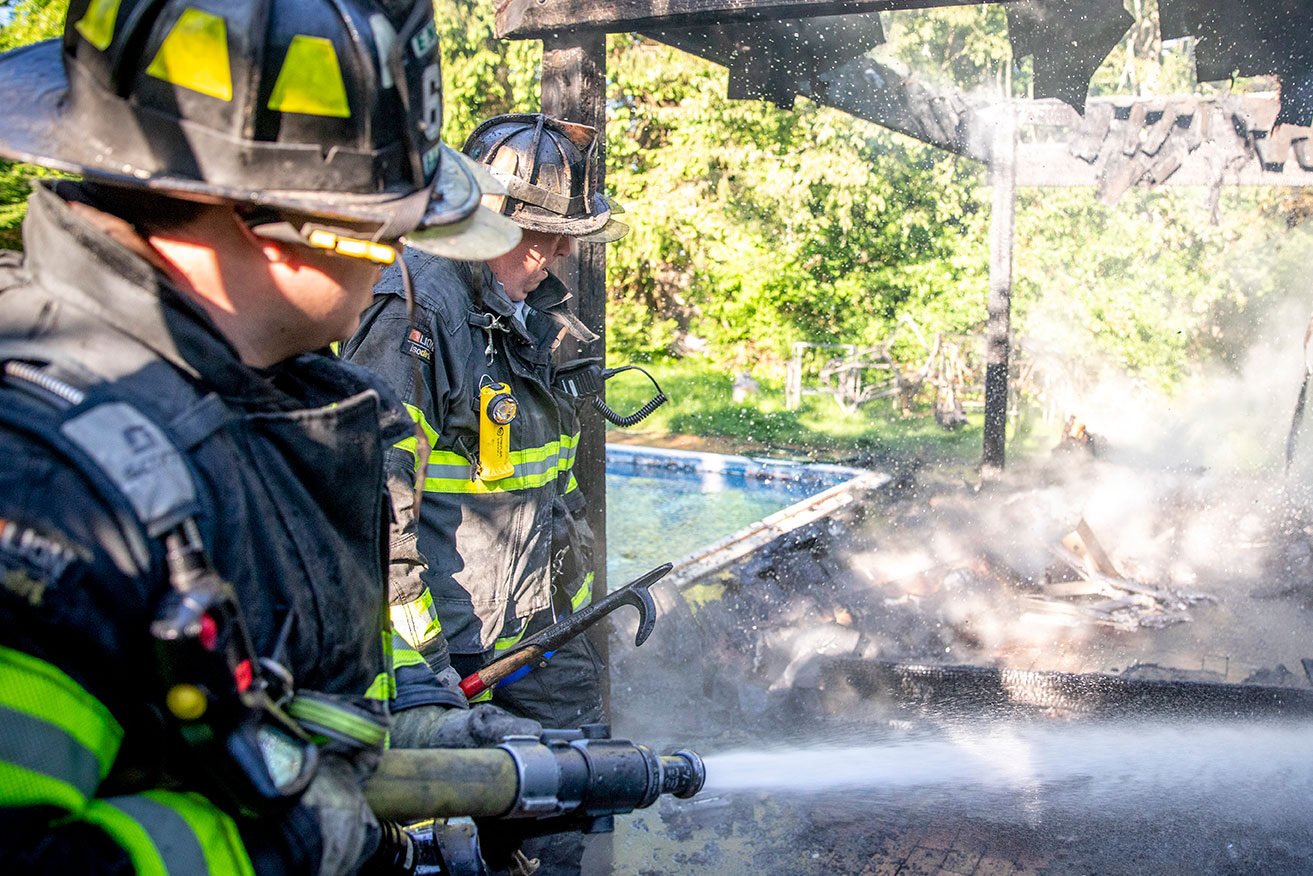 Two firefighters in turnout gear putting out a fire with a fire hose on a patio beside a swimming pool.