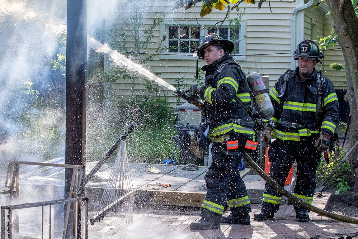 Two firefighters in turnout gear spraying water with a fire hose onto a patio to put out a fire.