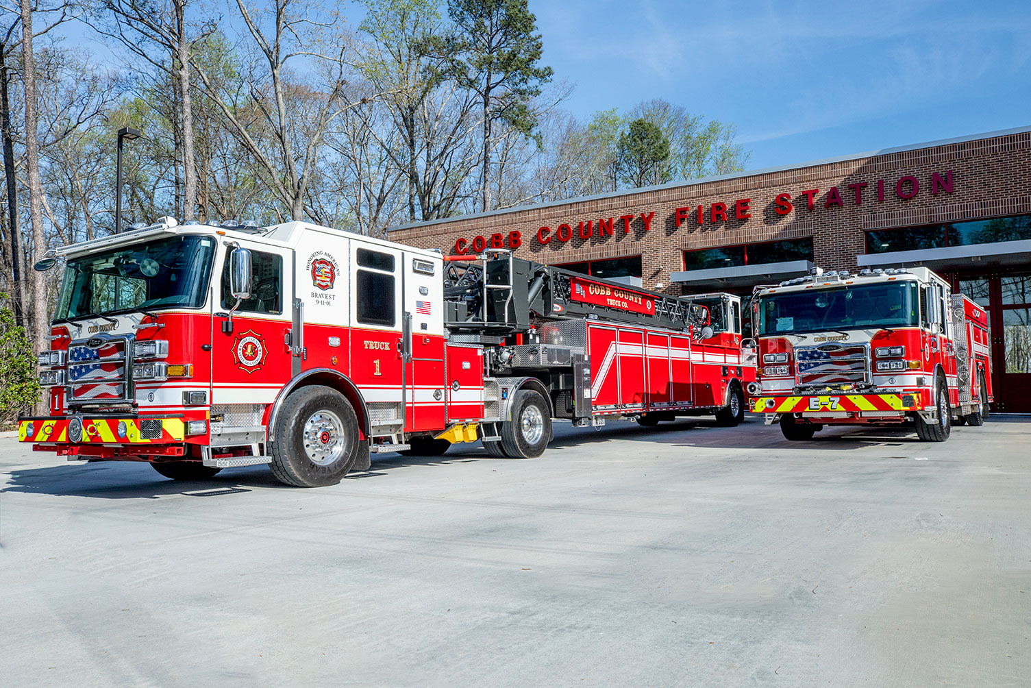 The front drivers side of a tractor drawn aerial and pumper pulling out of a fire station on a sunny day.