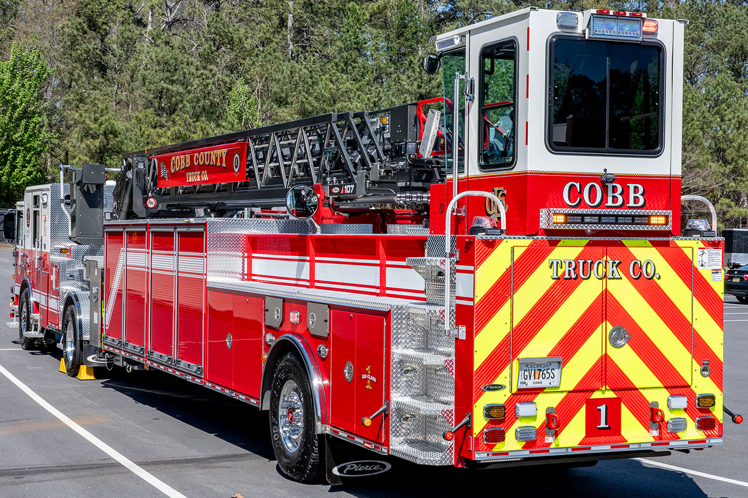 The rear drivers side of a tractor drawn aerial in a parking lot with the wheel chocks down by a treeline.