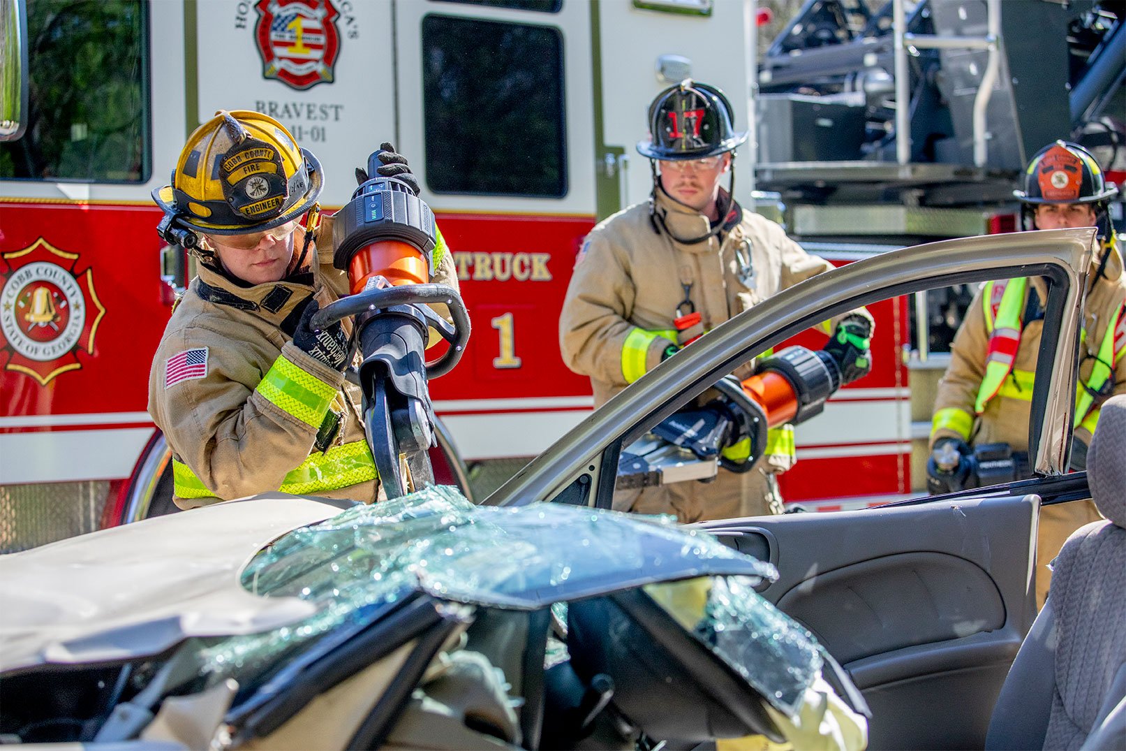 Three firefighters in turnout gear in front of a fire truck working with extrication equipment on a car.