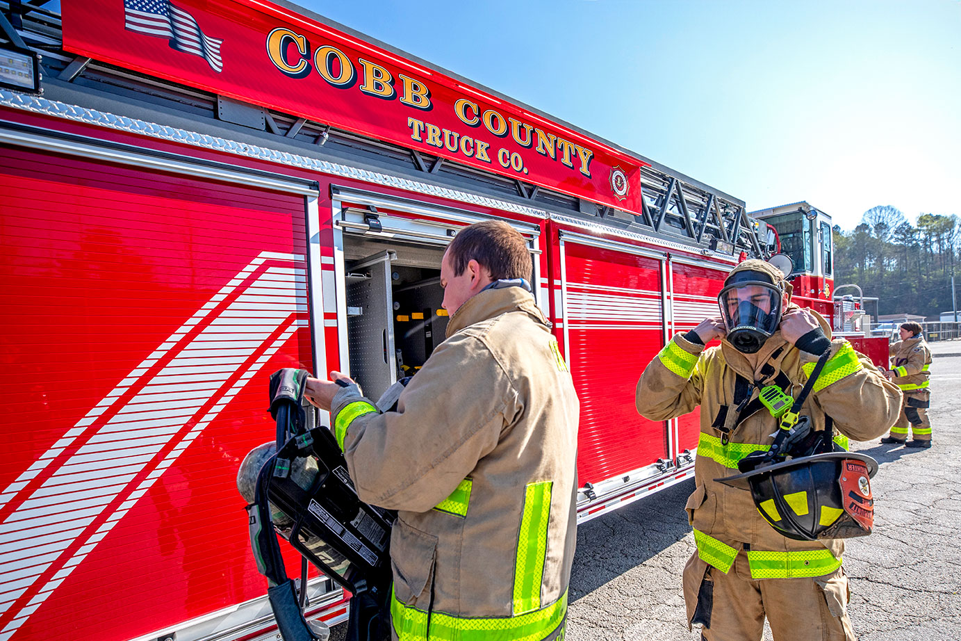 Two firefighters in turnout gear putting on SCBAs and masks beside a tractor drawn aerial on a sunny day.