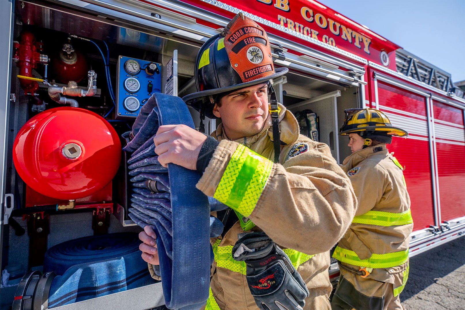 Two firefighters in turnout gear working off the side of a tractor drawn aerial grabbing hose and equipment.