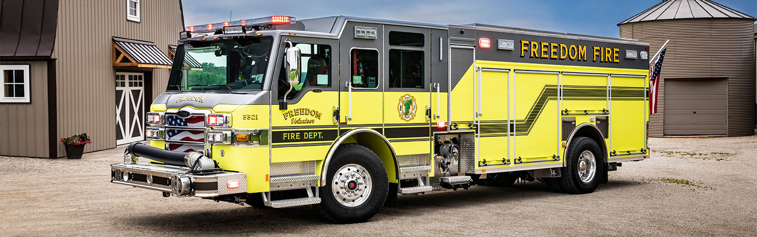 The front driver's side of a Velocity PUC Pumper parked by a barn on gravel on a partly cloudy day.